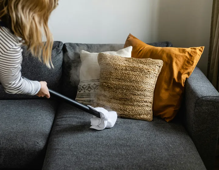 A steam wand with a white cloth lifting dust from a grey sofa cushion