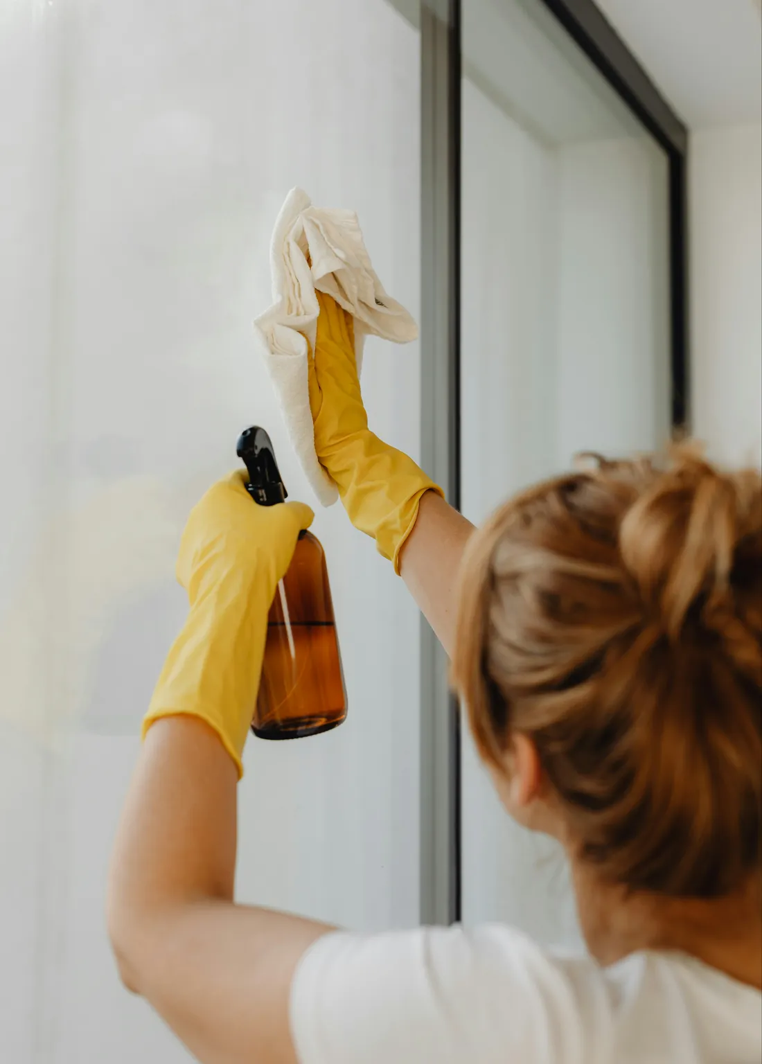 A cleaner in yellow gloves wiping a tall window with a cloth, a spray bottle in her other hand