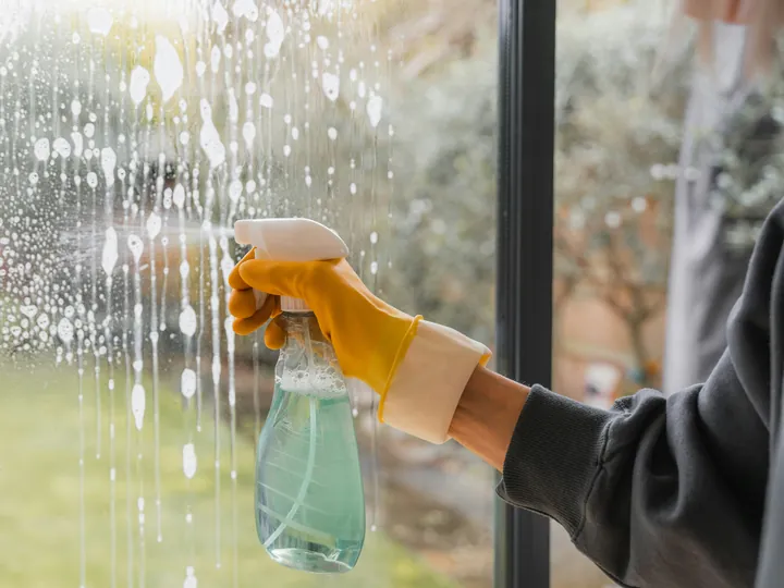 A gloved hand spraying cleaner on a garden window, suds running down the glass