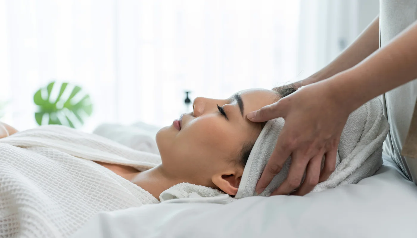 An esthetician's hands resting on a client's temples at the end of a facial, daylight through white curtains