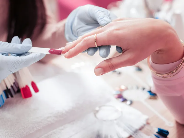 A nail tech in blue gloves holds a berry colour swatch against a client&rsquo;s nail