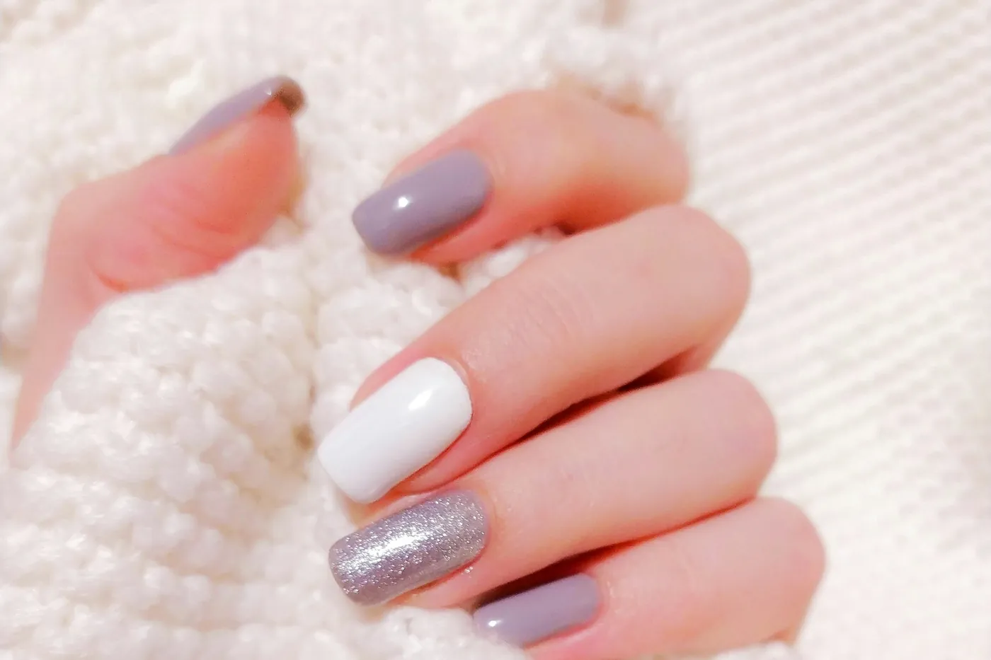 A hand resting in a cream knit sweater, short square nails in soft mauve with one milk-white nail and one silver shimmer nail