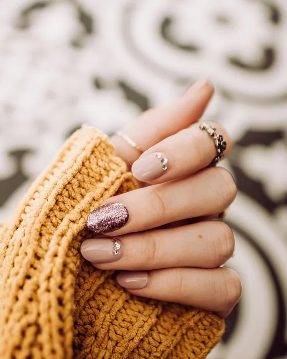A hand in a mustard knit sleeve, short oval nude nails with one rose glitter nail and two small crystals