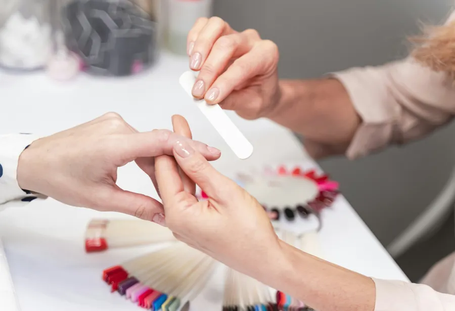 A tech files a client&rsquo;s short natural nail at a white table with colour swatch wheels nearby