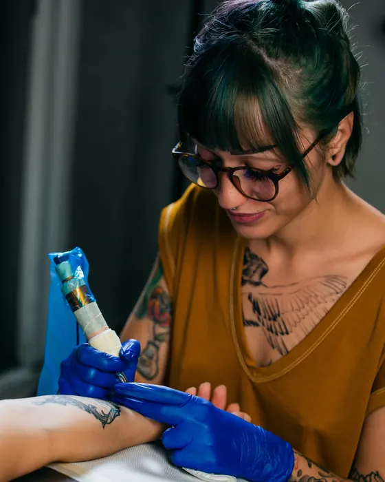 Mira, in glasses, tattooing a small design on a client's forearm